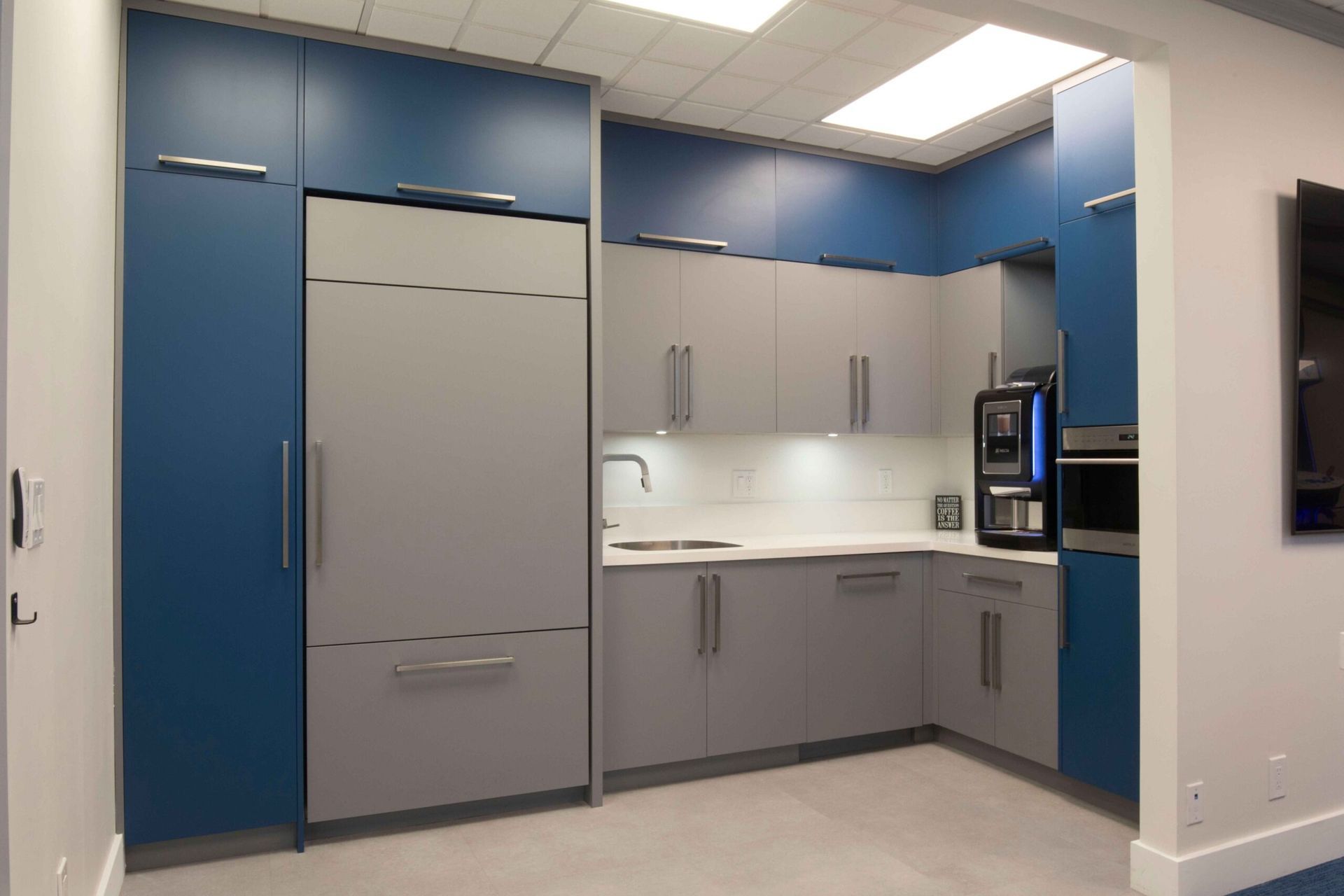 Modern office kitchenette with blue cabinets, gray counters, refrigerator, sink, and built-in microwave wall oven