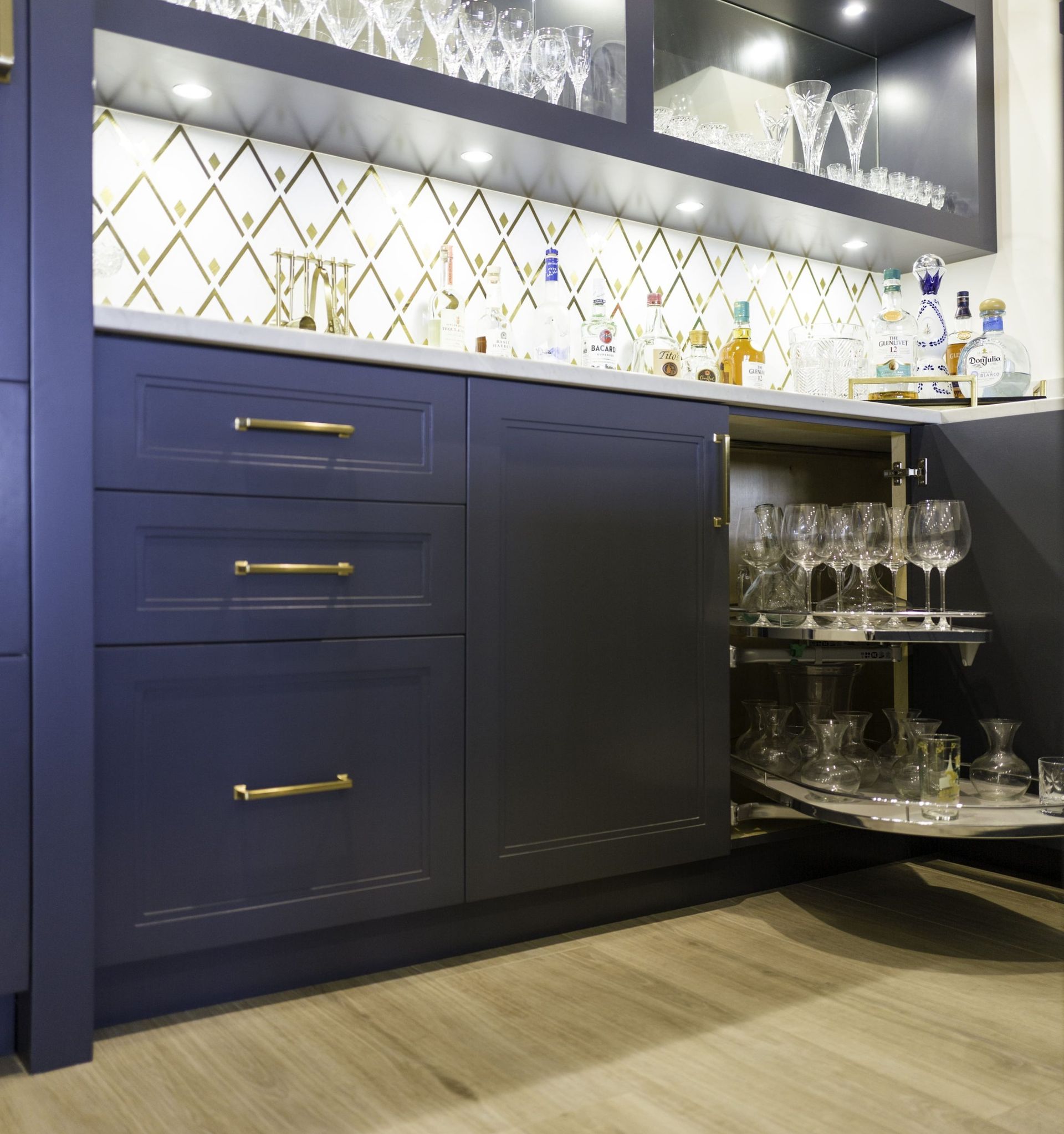 Blue kitchen cabinets with gold handles, lit mosaic backsplash, and open glass shelving with dishes