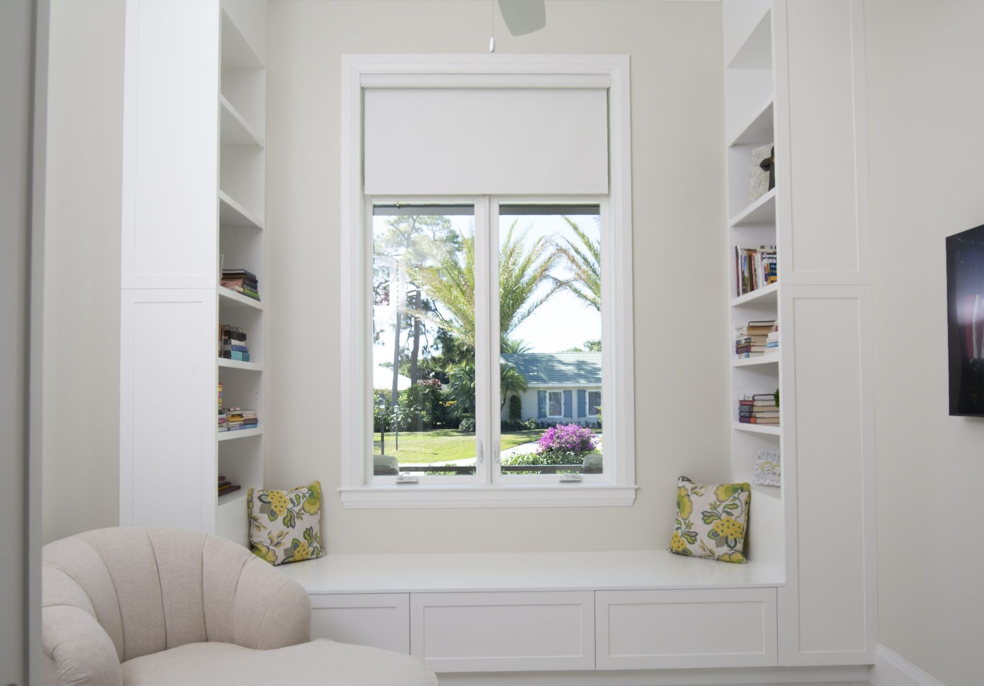Bright white window seat with built-in shelves, cushions, and a view of palm trees and flowers outside