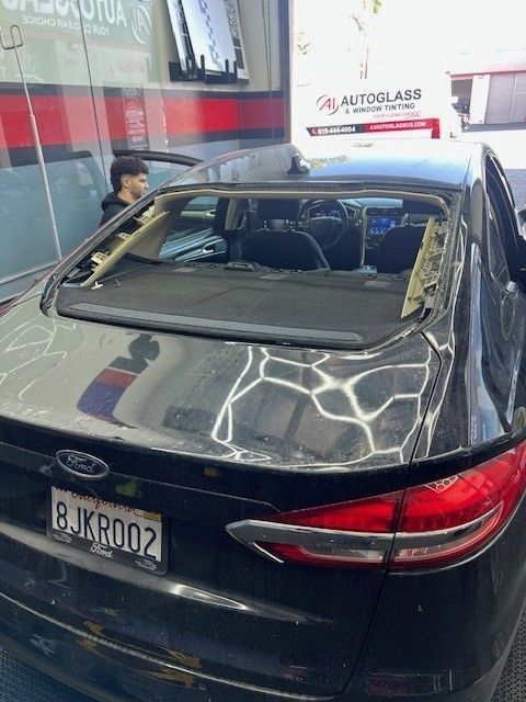 A black Ford Fusion with the rear windshield missing, parked in a car glass repair shop. A person stands nearby.