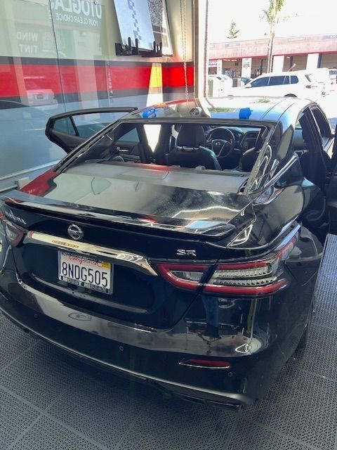 Black Nissan Maxima with a broken rear window at an auto shop. Doors open, and a person is visible inside.