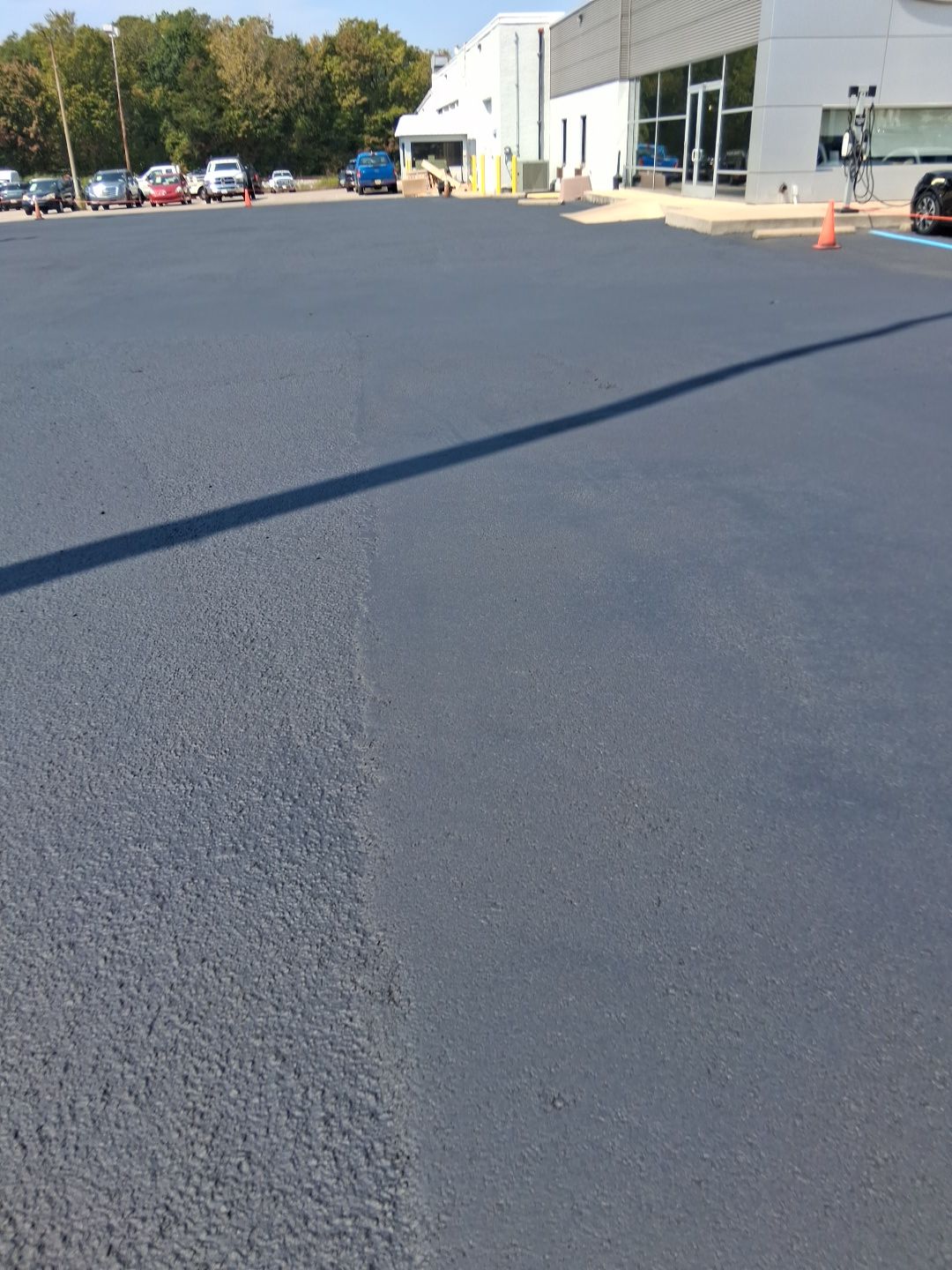 Freshly paved dark asphalt parking lot with cars and a building in the background.