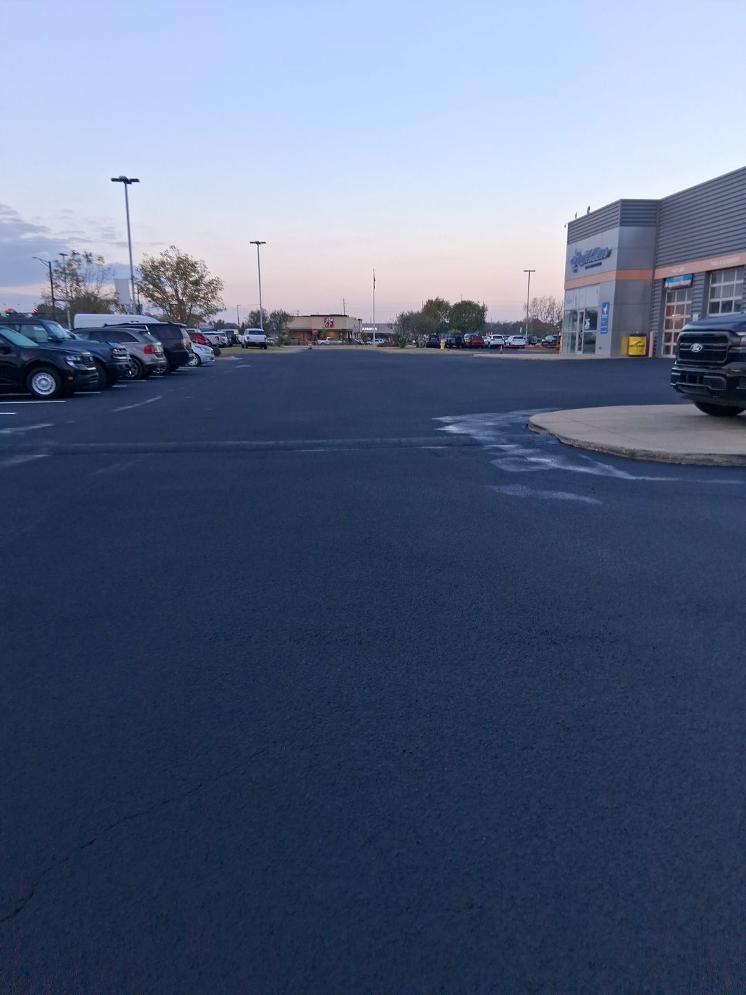 Empty parking lot in front of a building with parked cars on the left and right sides under a dusky sky.