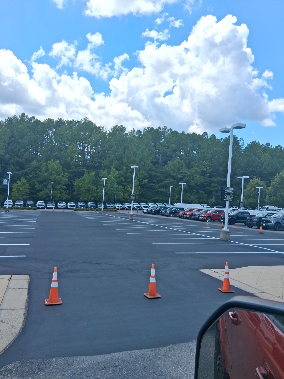 Car dealership parking lot on a sunny day with blue sky and white clouds; orange cones in the foreground.