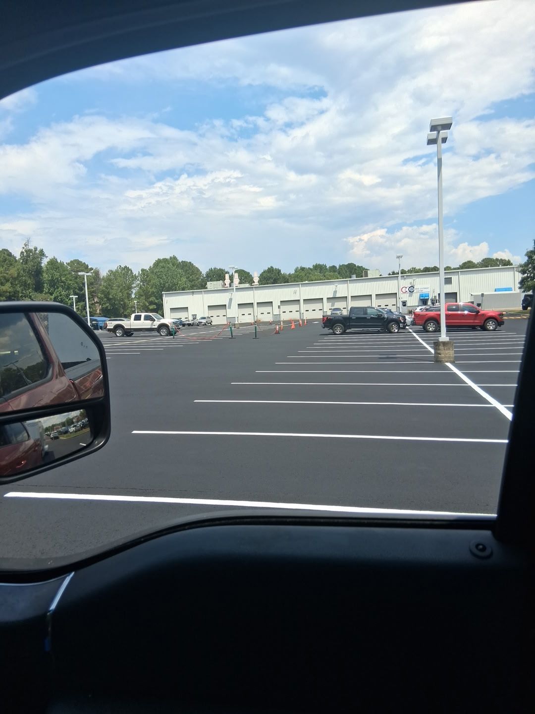 View from a car window of a mostly empty parking lot with a dealership in the background on a sunny day.
