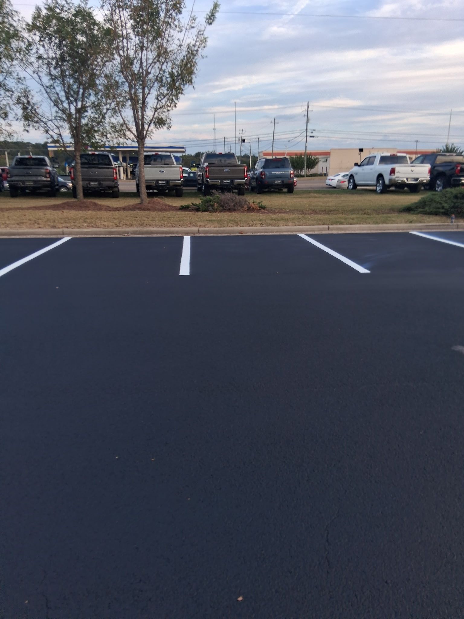 Empty parking spaces in the foreground; several parked trucks in the background. Blue sky and some trees visible.