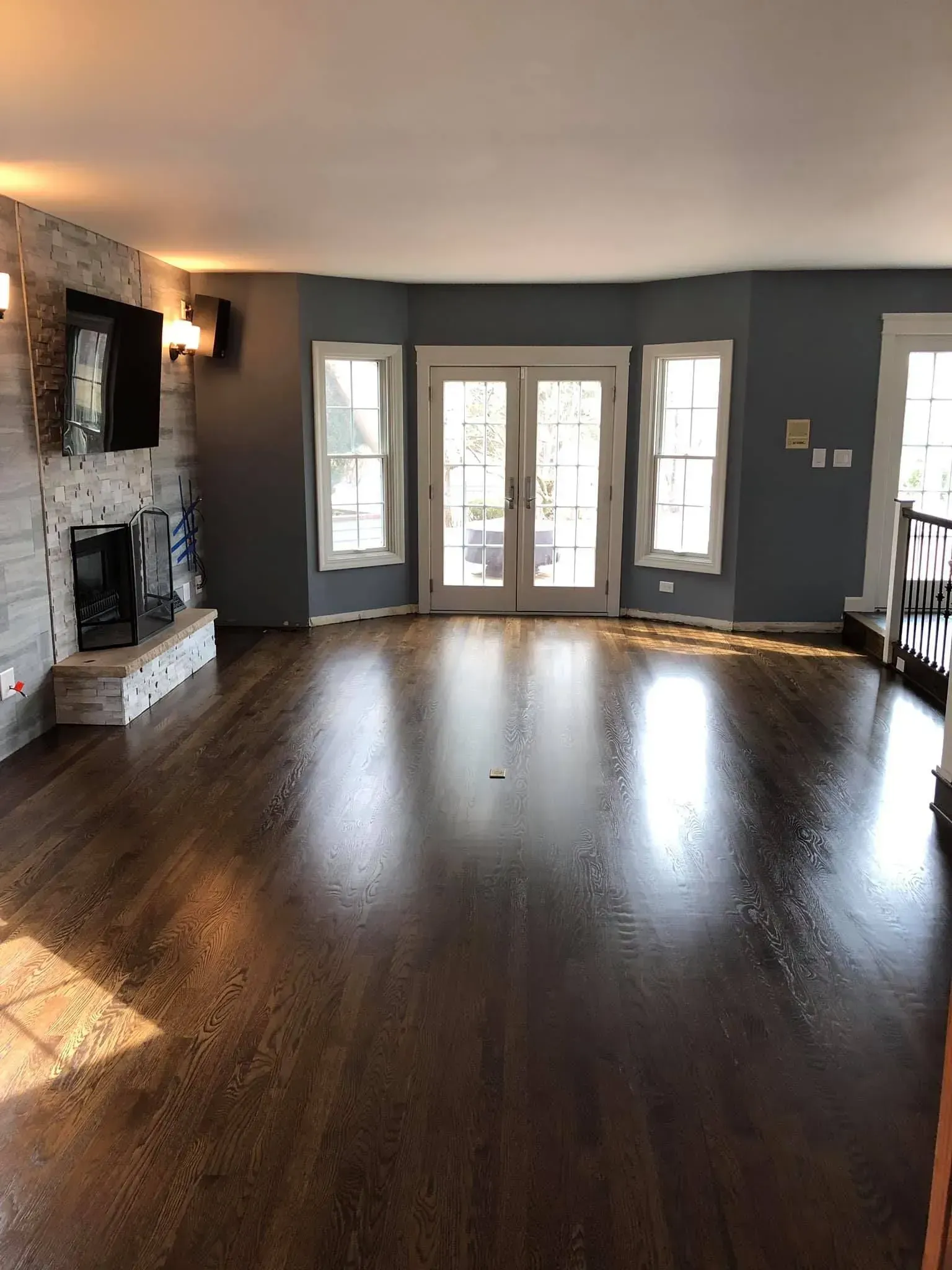 Empty living room with dark wood floors, gray walls, stone fireplace, and French doors to a sunlit patio