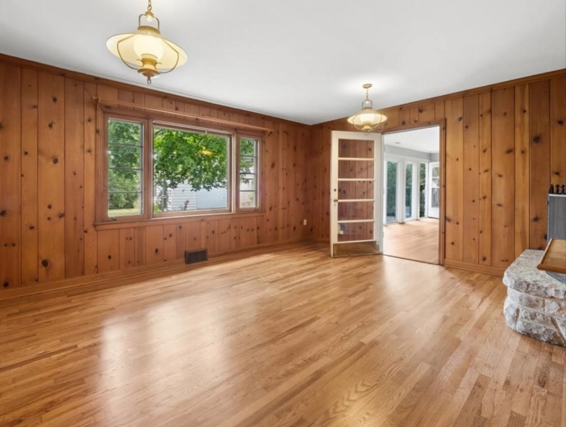 Wood-paneled empty living room with hardwood floors, windows, and a doorway to a bright sunroom.