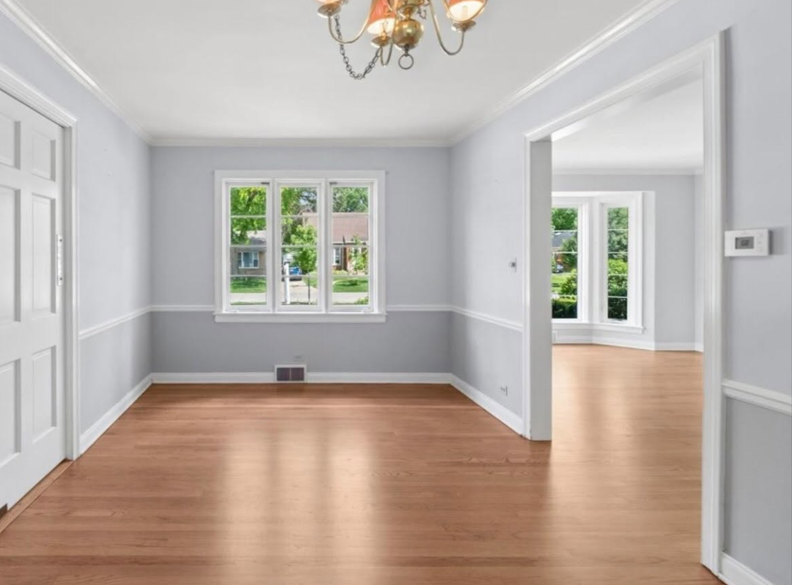 Empty light-gray room with hardwood floors, white trim, and windows overlooking greenery.