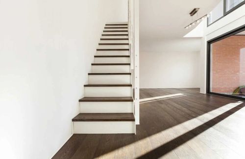 Modern empty interior with staircase, dark wood floor, and large window casting sunlight across the room