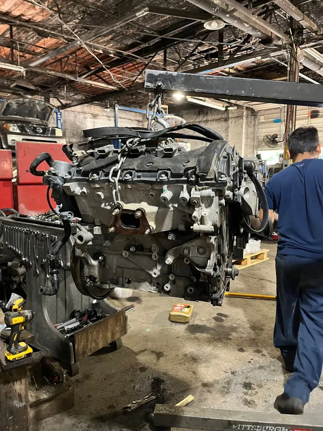 An engine suspended by a crane in a repair shop, mechanic in blue shirt beside it.