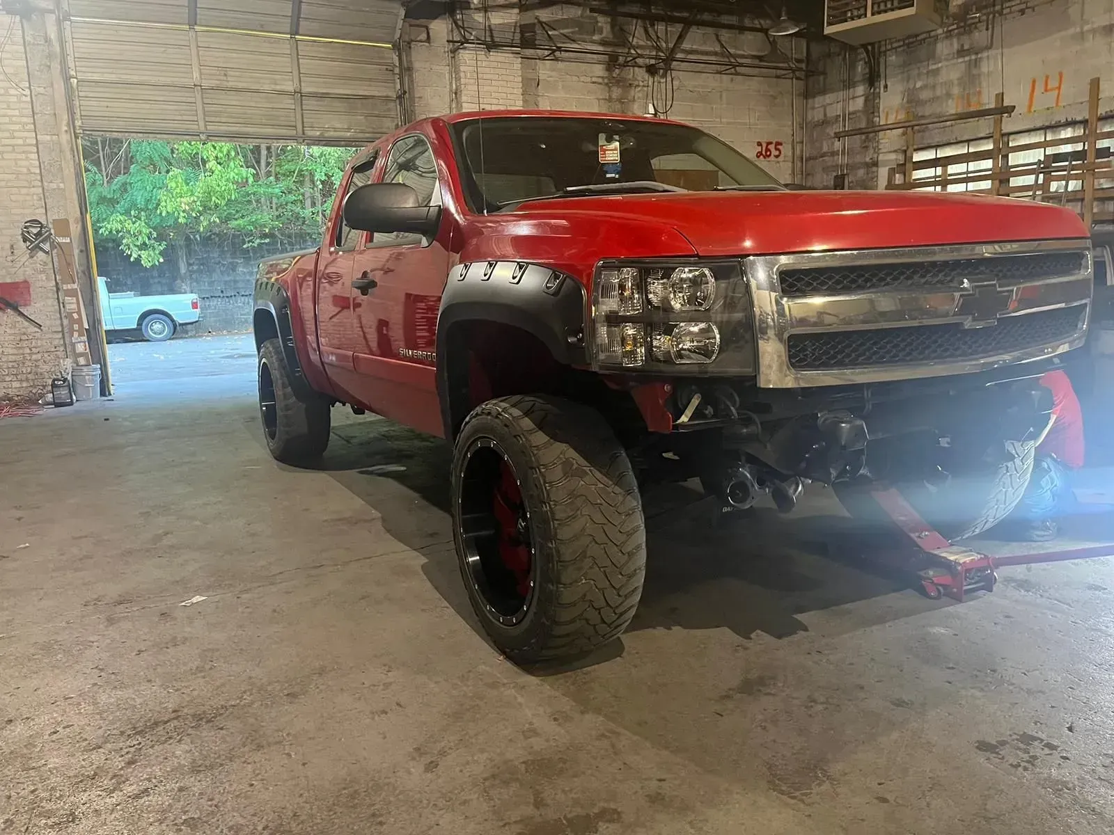 Red Chevrolet truck in a garage; front end damaged, black rims, gray walls.