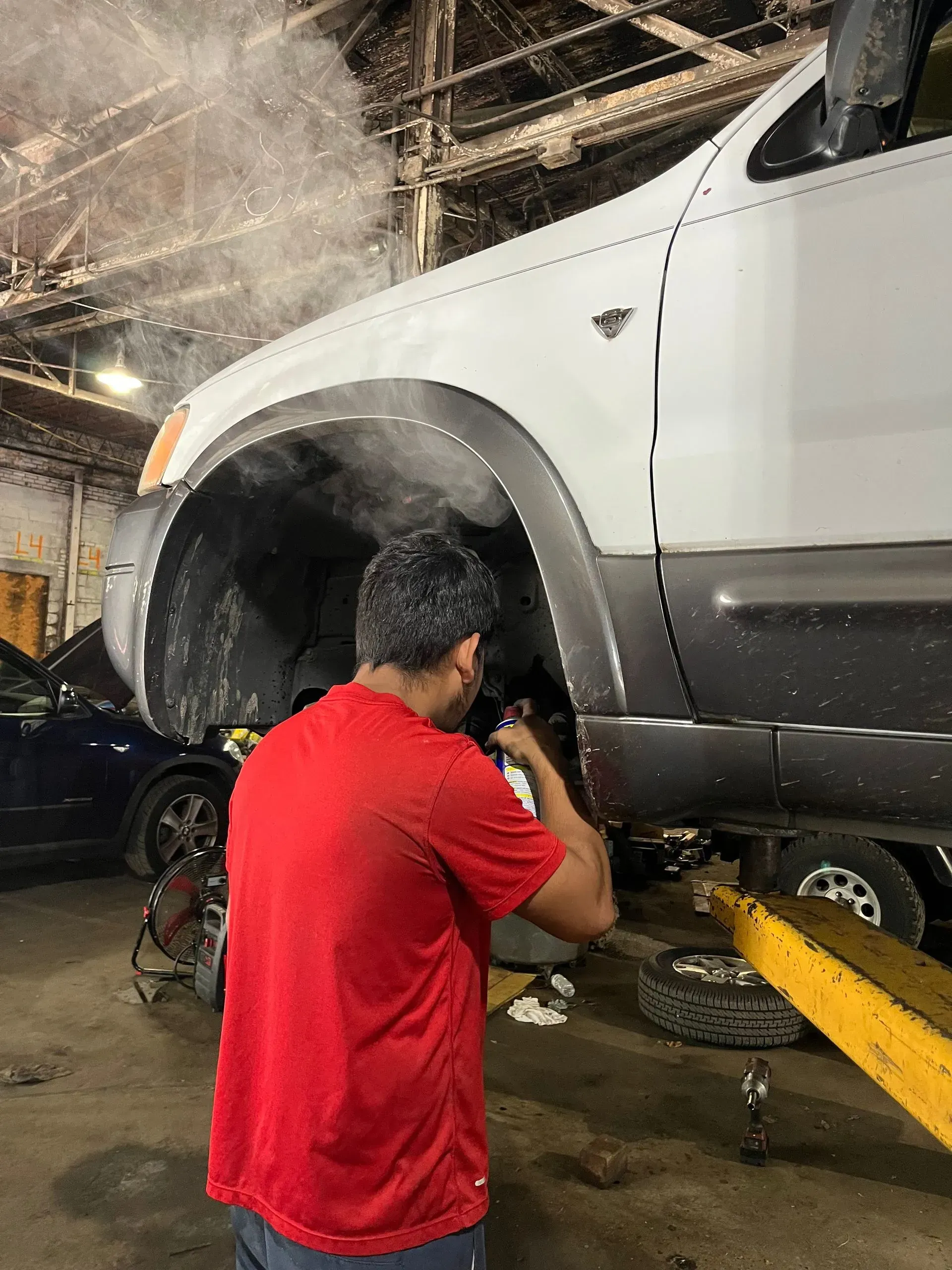 Mechanic working on a raised white car in a garage, steam rising from the wheel well.
