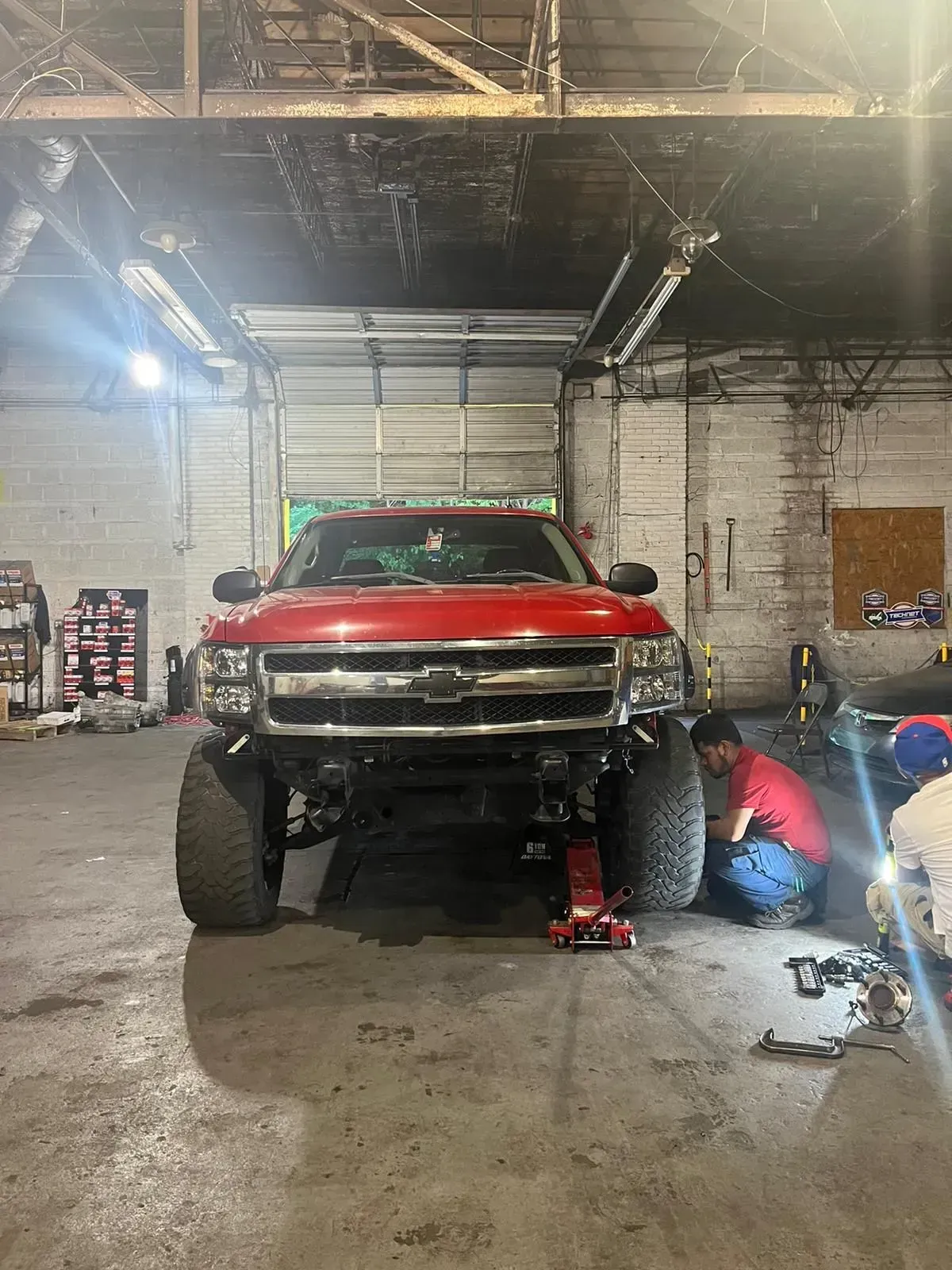 Red truck being worked on in a garage; a person kneels, red jack, large tires.