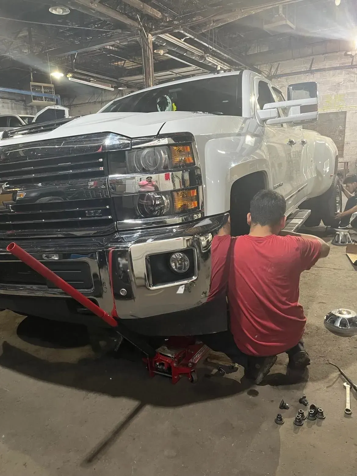 Person working on the front of a white truck in a garage; red jack is used.