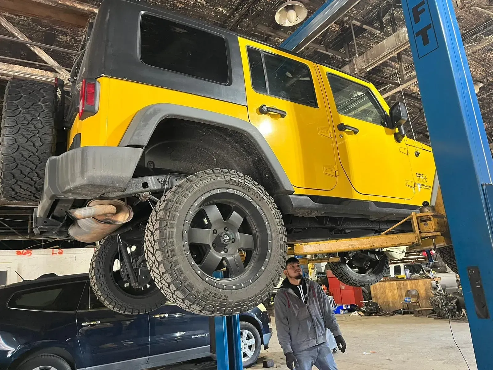 Yellow Jeep Wrangler on a lift in a repair shop. A person stands nearby.