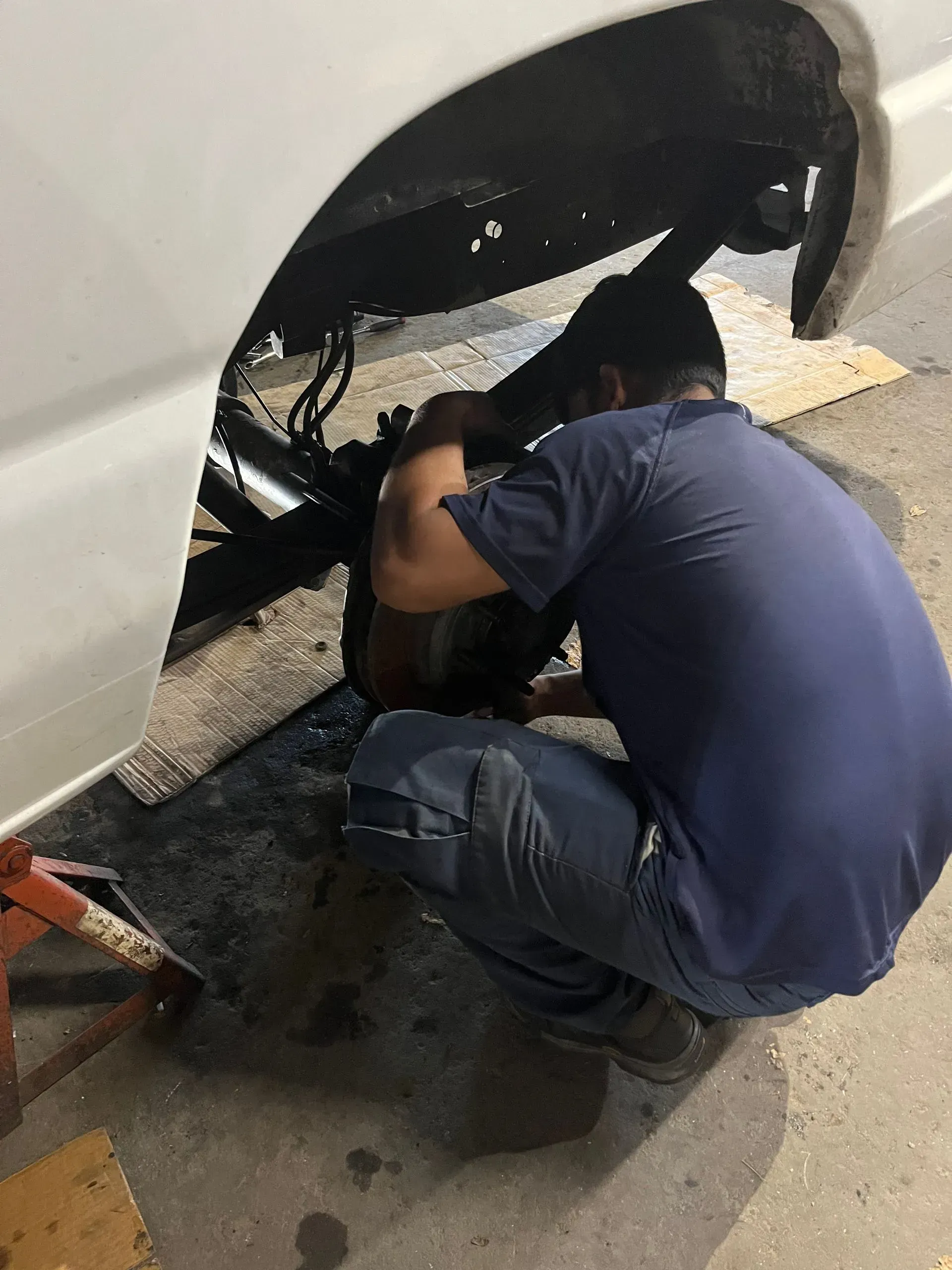 Mechanic working on a car's wheel, crouching near the tire. White car, dark setting, and a jack stand.