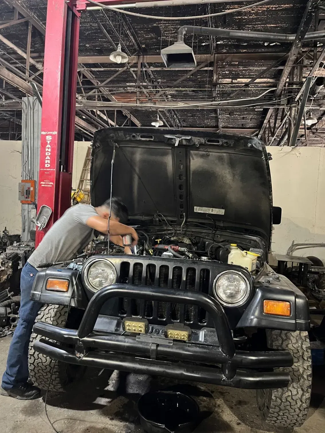 Mechanic working on black Jeep with hood open in a garage, red lift on left.