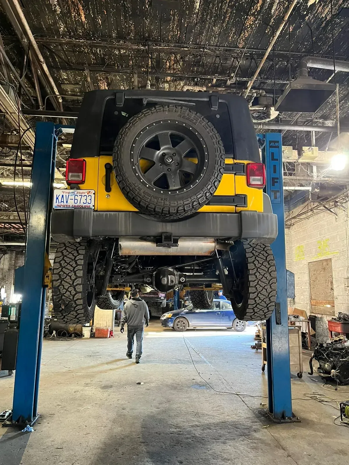 Yellow Jeep Wrangler raised on a lift in a garage. A person walks underneath.