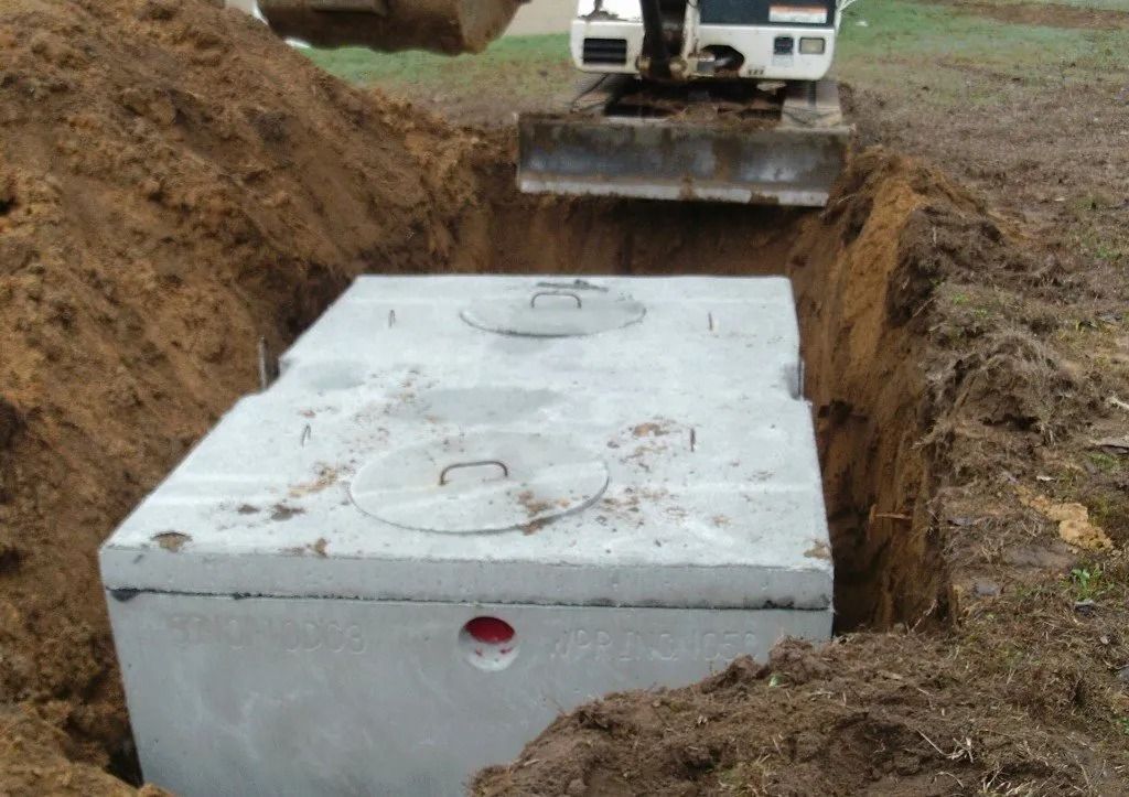 A concrete septic tank being lowered into a trench by a small excavator on a grassy area.