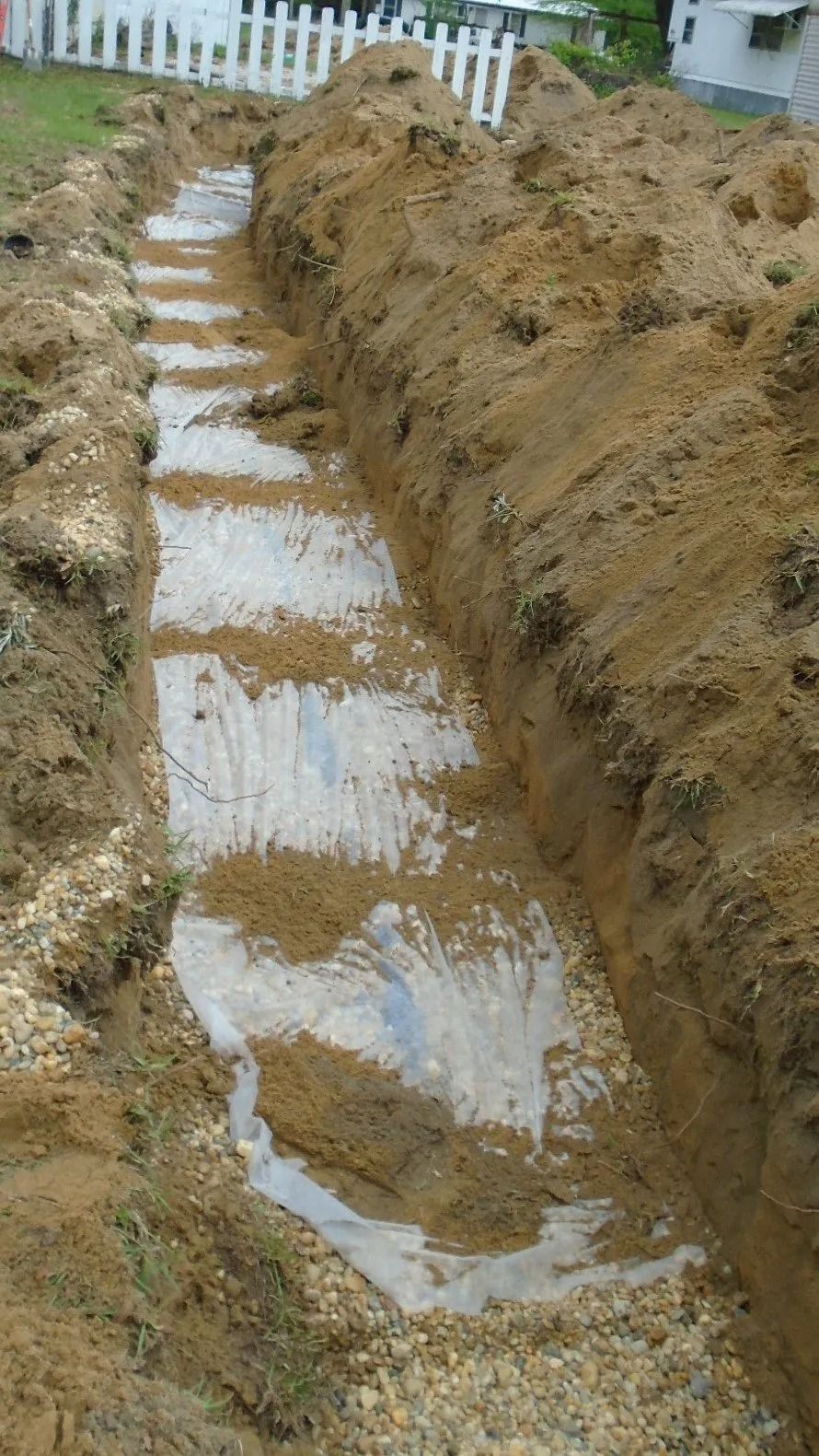Long, narrow trench in dirt with gravel and white cloth, near a white picket fence.