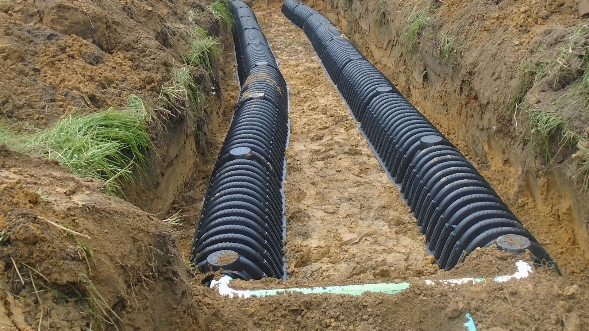 Two parallel corrugated plastic pipes in a trench, part of a drainage or septic system, surrounded by brown soil.