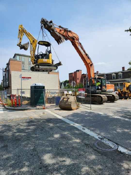 A yellow excavator is being lifted by a large orange excavator.