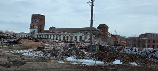 Demolition of a historic brick factory building under a cloudy sky, with debris in the foreground and graffiti on walls.