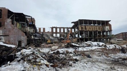 A partially demolished brick industrial building with debris and snow in the foreground under a cloudy sky.