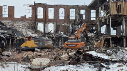 Two construction excavators work to demolish a large, multi-story brick building surrounded by piles of rubble and snow.