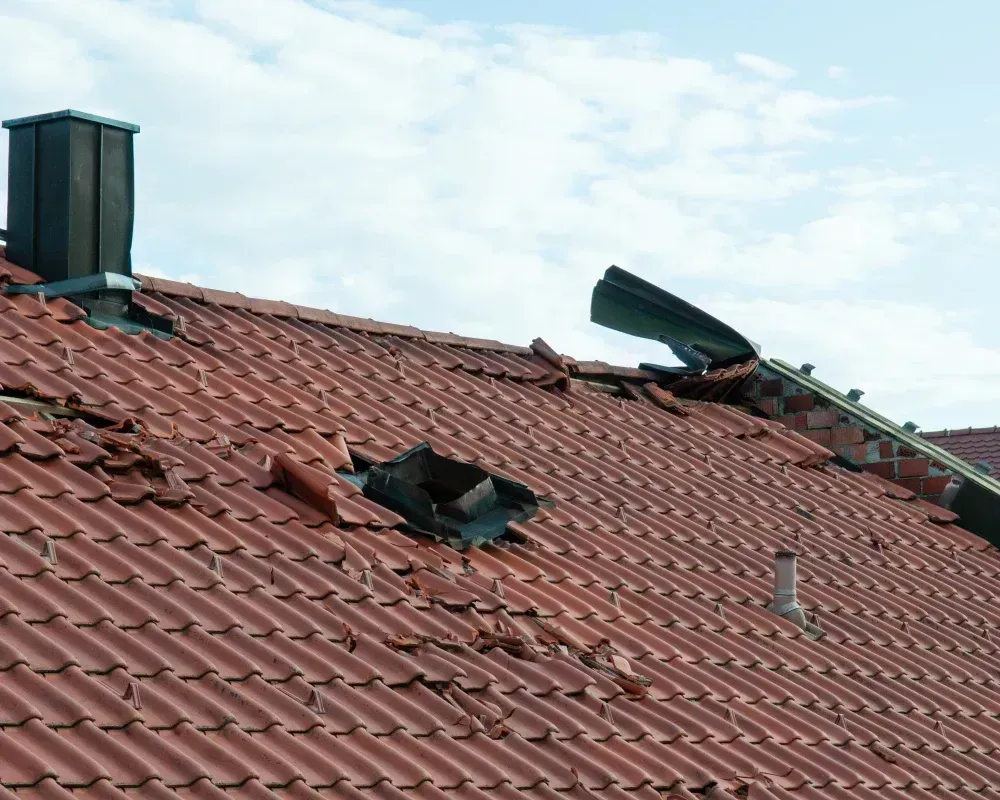 Damaged red tile roof with missing tiles and a bent chimney vent under a cloudy sky