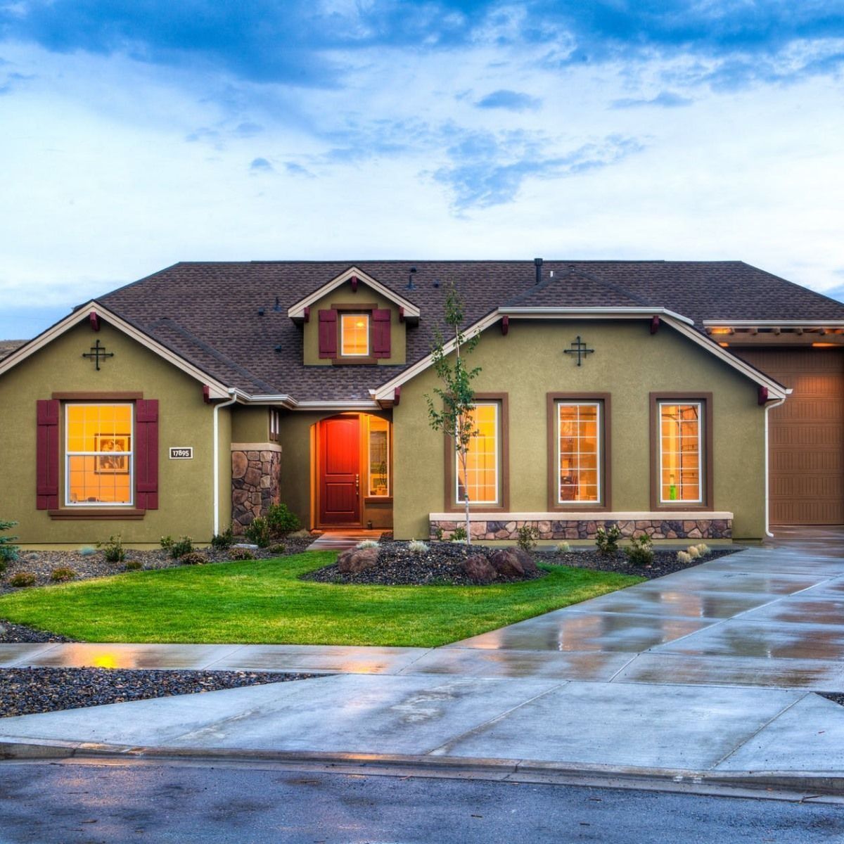 Modern single-story house with lit windows, a wet driveway, and a green lawn at dusk