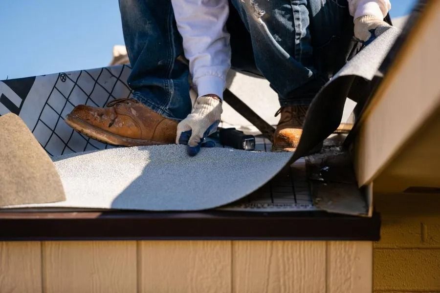 Worker installing roofing felt on a roof edge with tools and wooden siding below