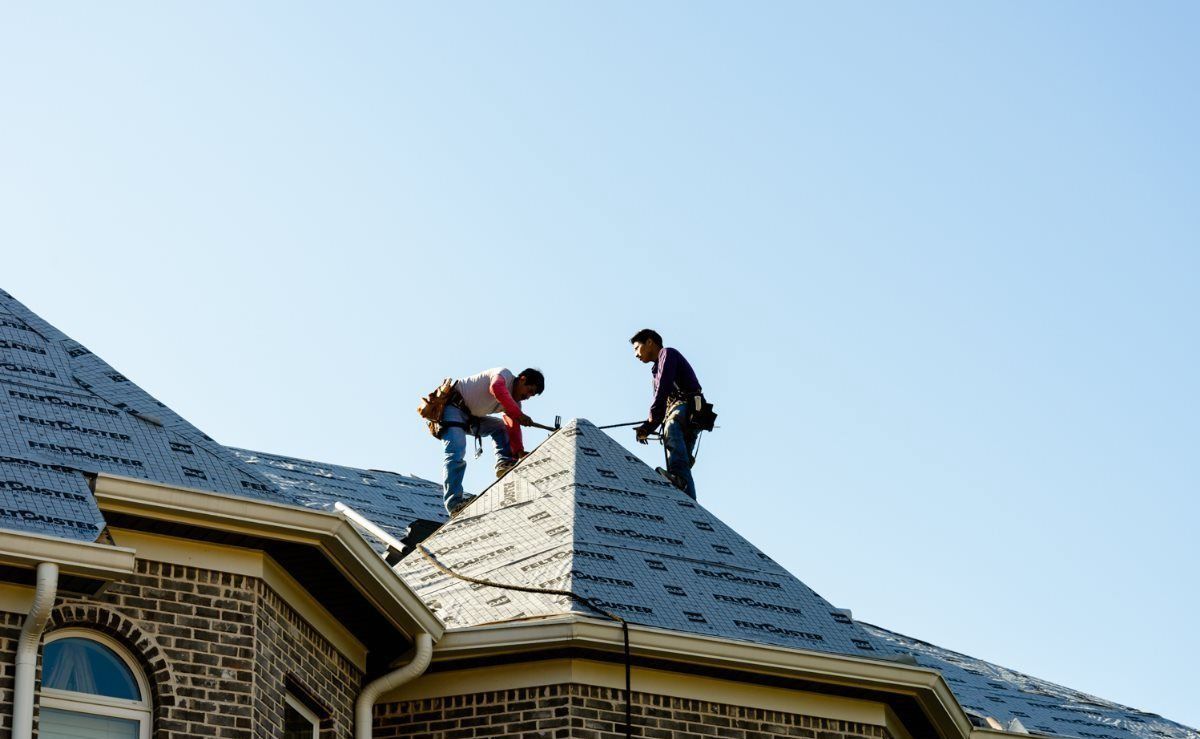 Two workers on a house roof, installing shingles under a clear blue sky