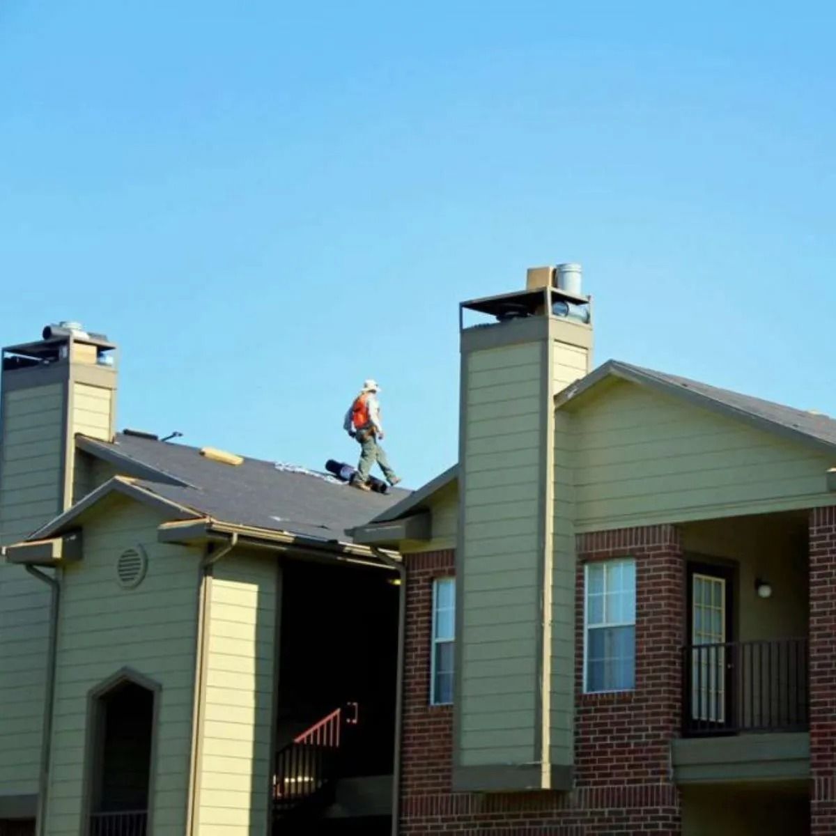 Worker on a roof repairing shingles on a townhouse under a clear blue sky