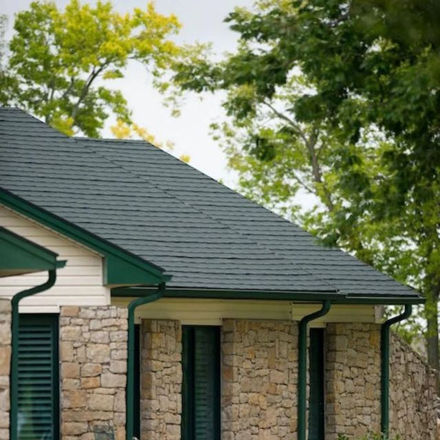 Stone house with green trim and dark roof beneath leafy trees