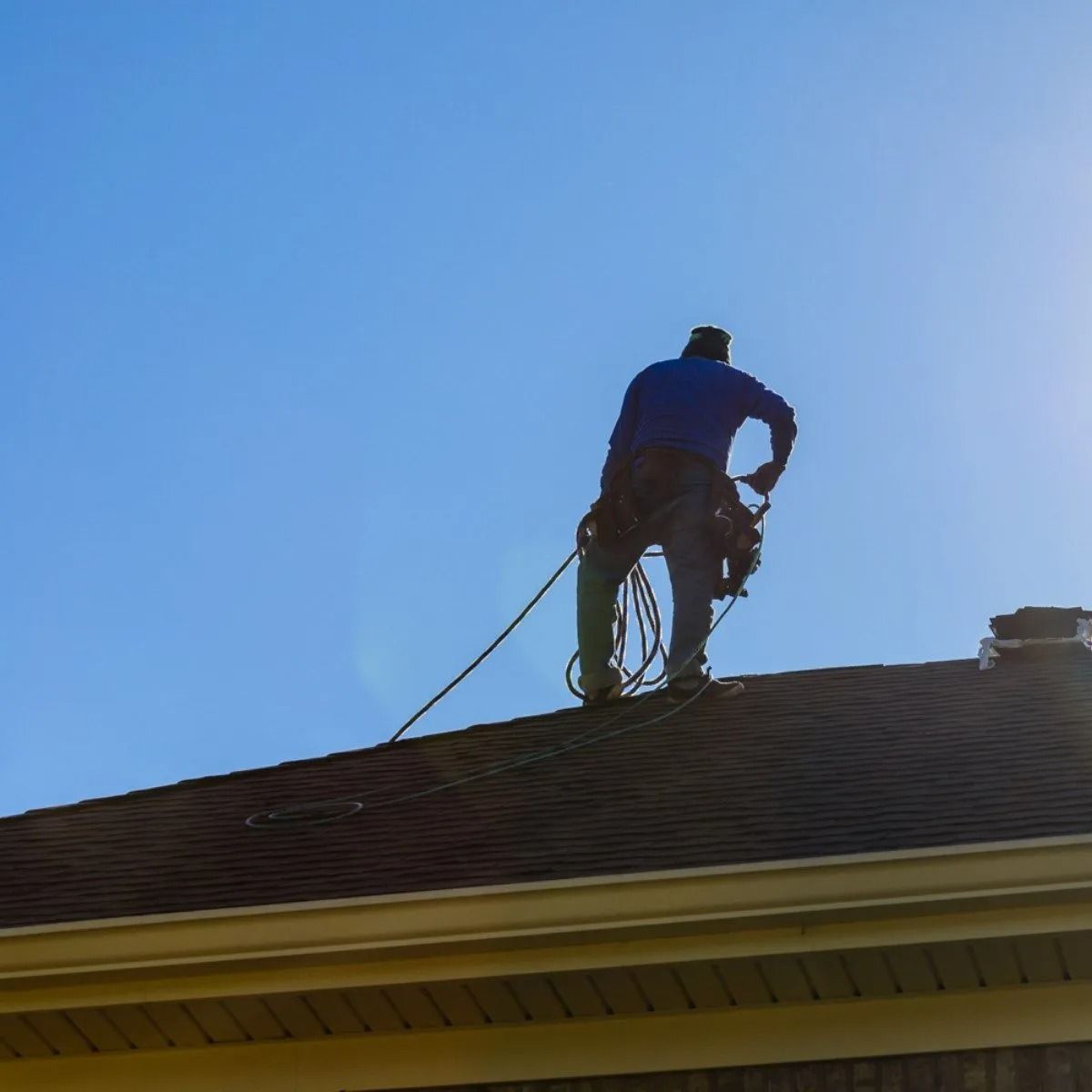 Worker on a rooftop repairing shingles under a clear blue sky