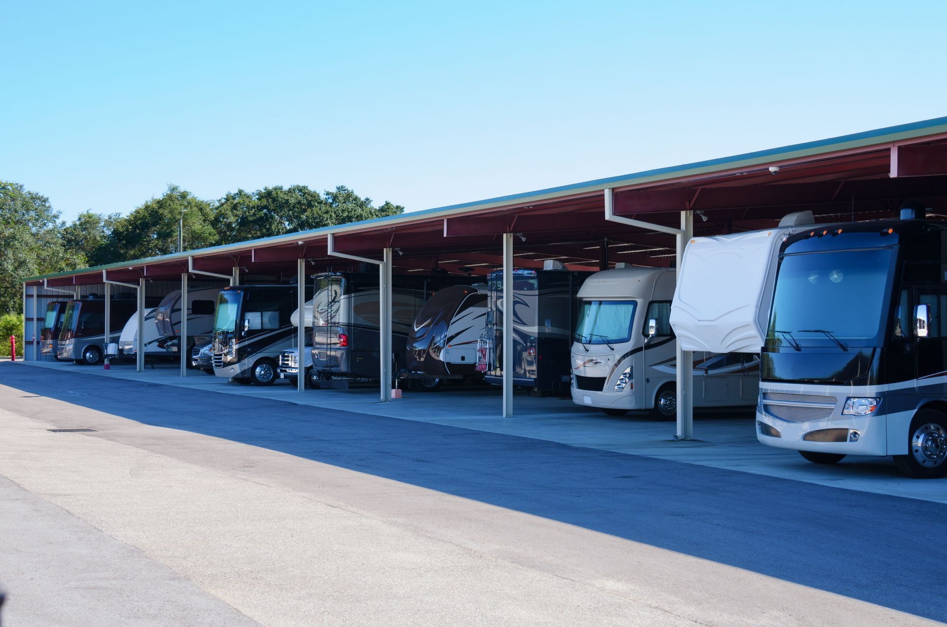 RVs parked under a long, covered shelter on an asphalt surface, under a blue sky.