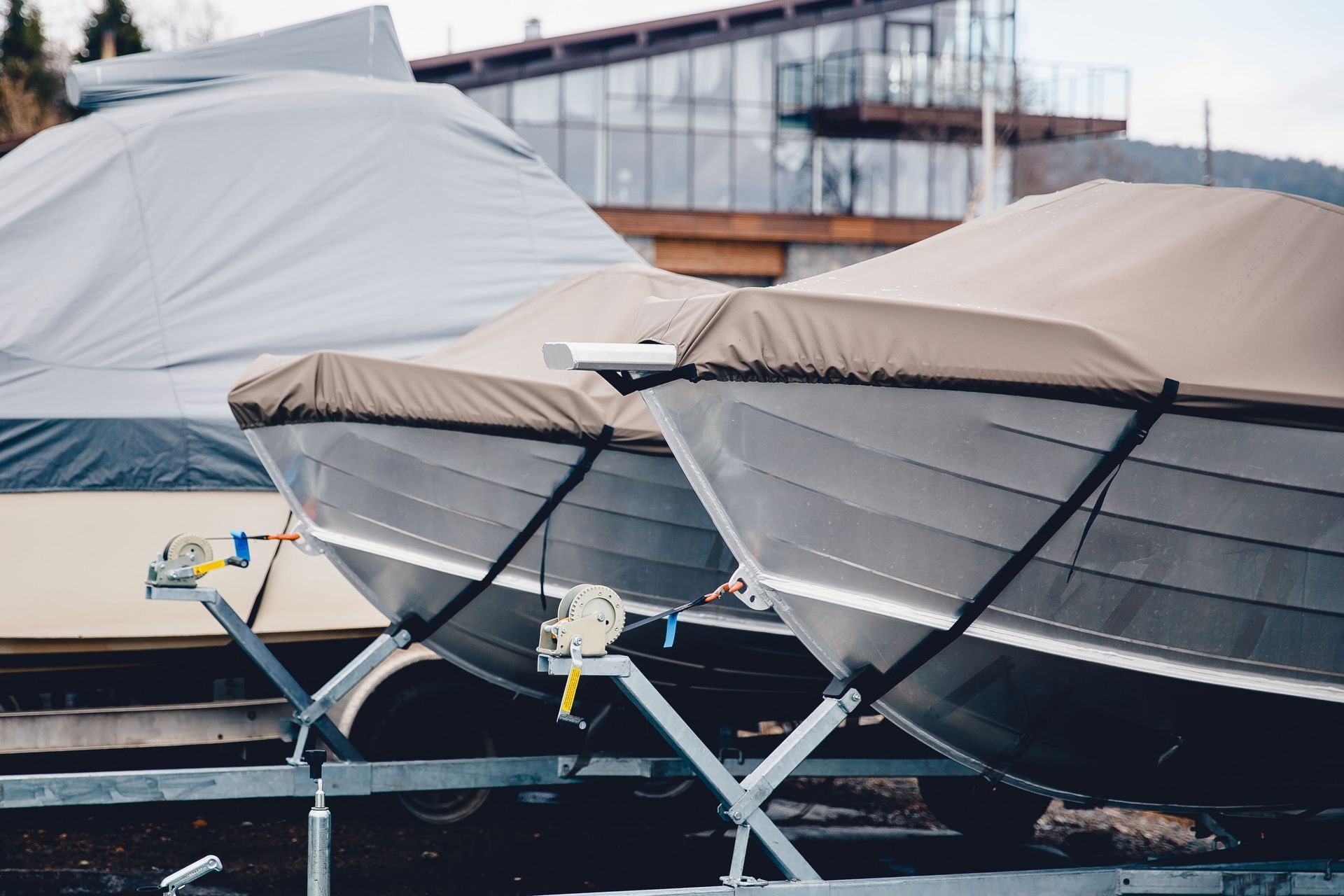 Boats on trailers, covered with gray and tan tarps; parked outside near a building.