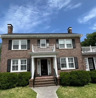 Brick house with black shutters, white trim, and a small balcony under a partly cloudy sky.