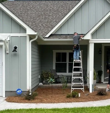 Man on ladder installing gutter on a light blue house with white trim.