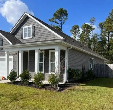 Gray house with a front porch, shutters, and landscaping, against a blue sky with trees in the background.