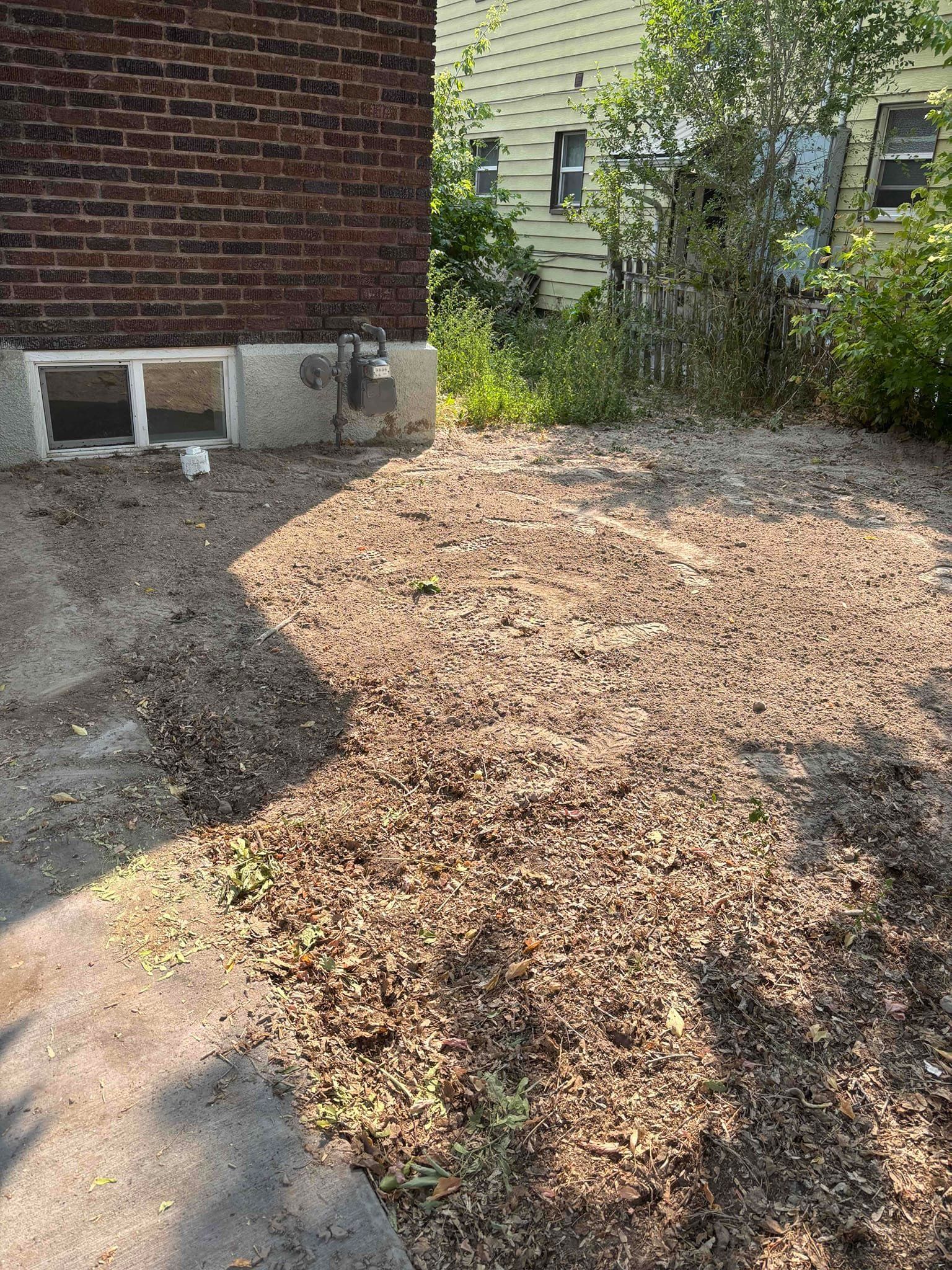 A gravel yard next to a brick building and a yellow house with overgrown bushes in the background.