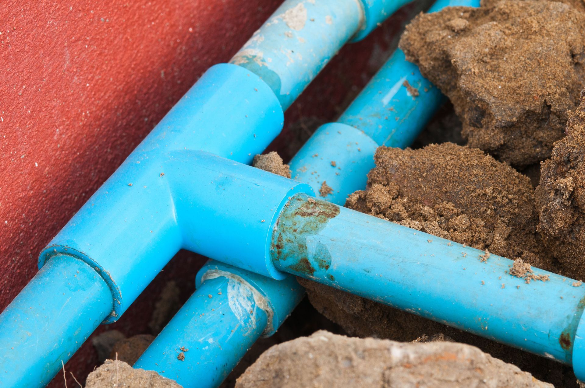 Blue PVC pipes with T-joints, partially buried in soil, next to a red surface.