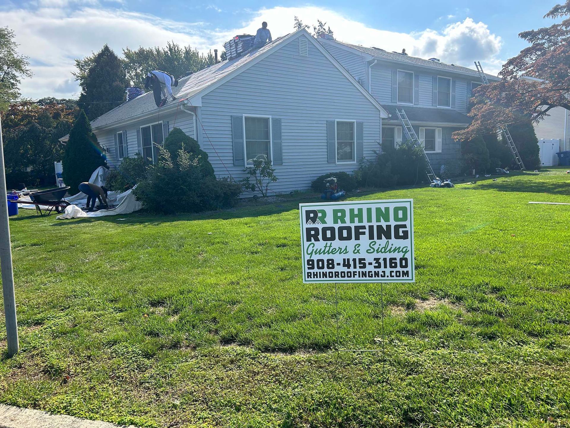 Roofers working on a house with a Rhino Roofing sign in the foreground, blue siding, green lawn.