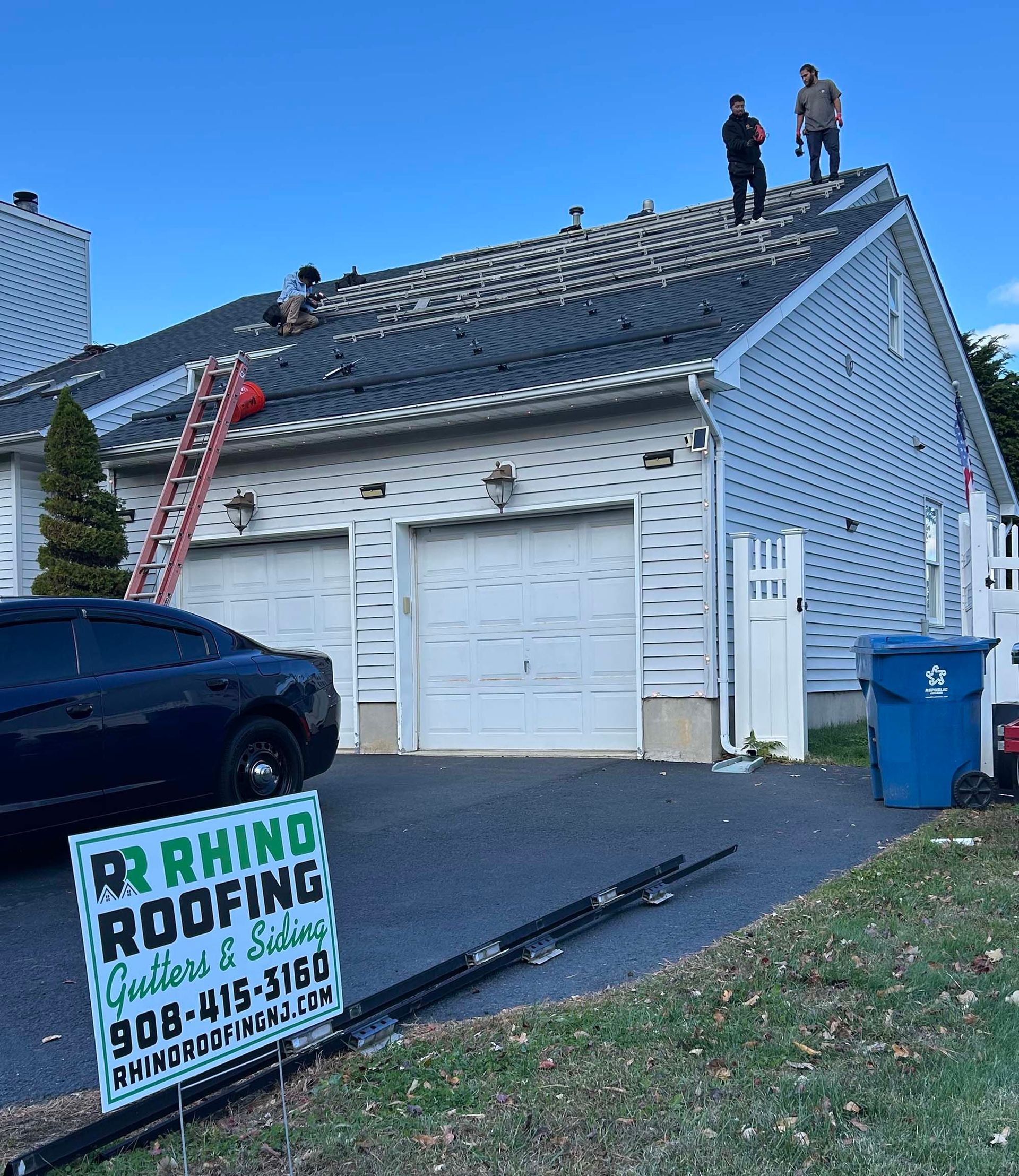 Roofers on a house roof replacing shingles. A ladder and a sign for Rhino Roofing are in view.