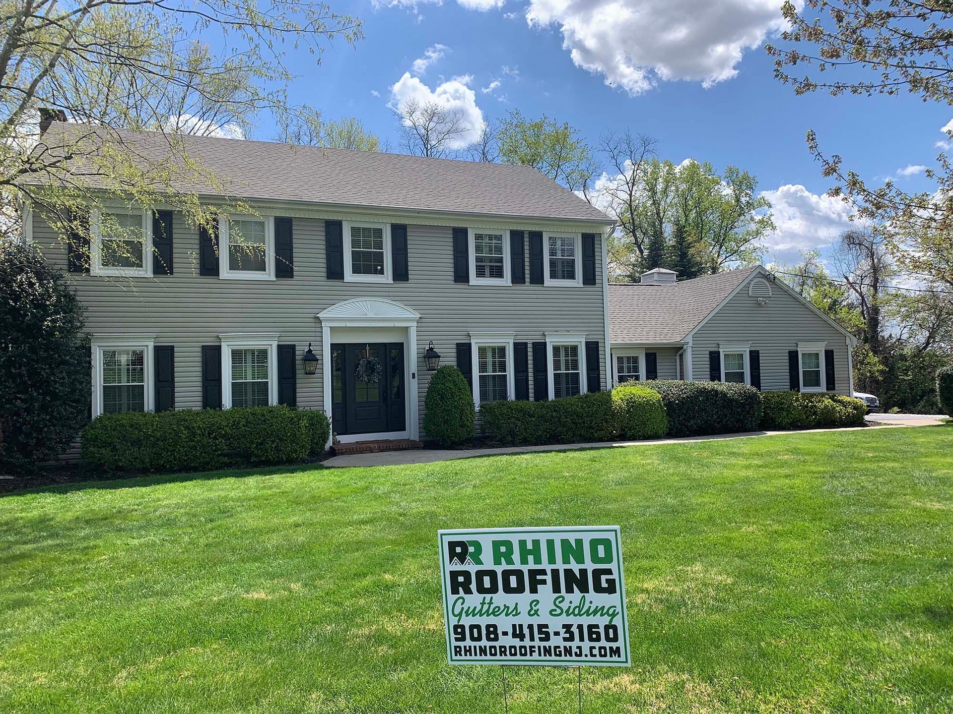 Two-story house with gray siding, black shutters, and a green lawn; a Rhino Roofing sign is in the yard.