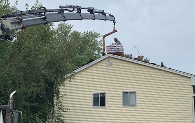A worker on a roof receiving materials from a crane. The house has tan siding and two windows.