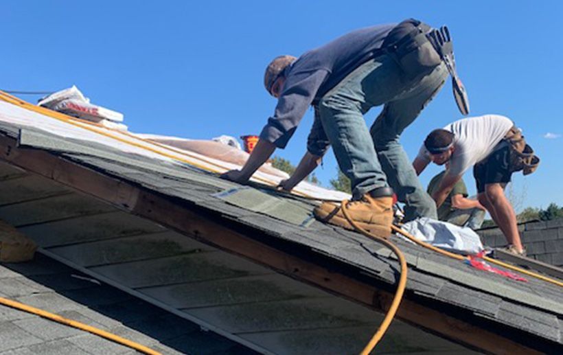 Three roofers installing shingles on a slanted roof under a blue sky; one kneeling, two bent over working.