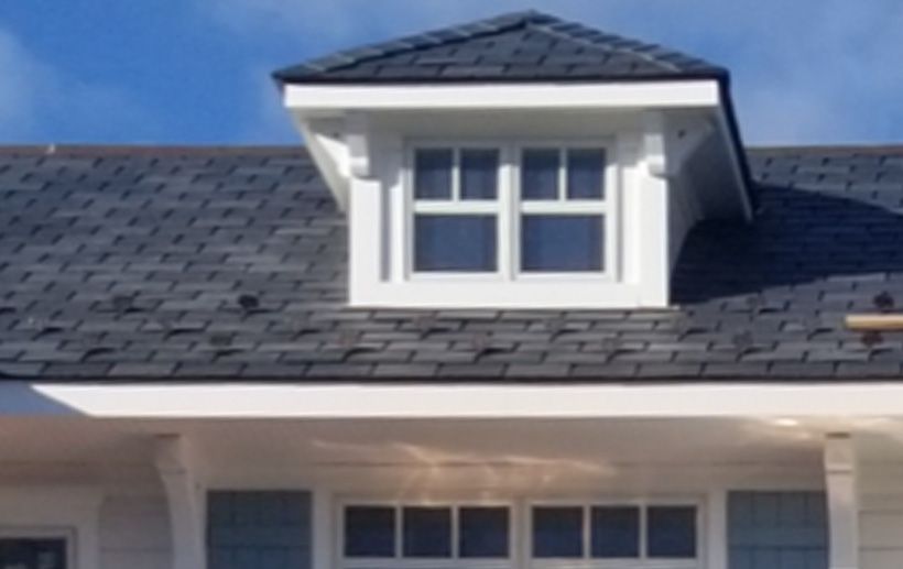Dormer with window on a gray shingled roof, white trim. Blue sky background.